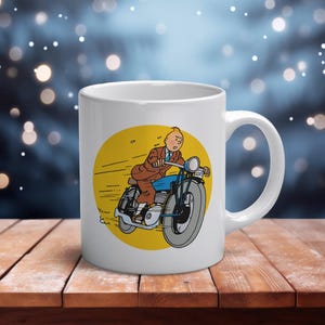 Puede incluir: Taza de cer&aacute;mica blanca con una ilustraci&oacute;n de dibujos animados de un hombre en motocicleta. La imagen est&aacute; sobre un c&iacute;rculo amarillo. El hombre lleva un traje marr&oacute;n y pantalones azules. La taza tiene un asa curvada.