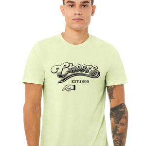 May include: A light green t-shirt with a black graphic that says "Cheers EST.1895" with a black hand pointing to the right.