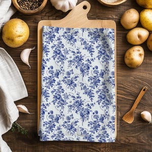 May include: A white and blue floral patterned kitchen towel. The design features blue flowers and foliage on a white background. The towel is folded on a wooden cutting board, surrounded by potatoes, garlic, and spices.