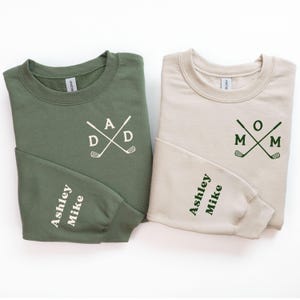 May include: Two crewneck sweatshirts, one olive green and one beige. The green sweatshirt has "DAD" with crossed golf clubs, and the beige one says "MOM". Both have "Ashley Mike" on the sleeves.