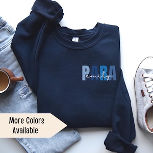 K&ouml;nnte beinhalten: Ein marineblauer Pullover mit dem Text "PARA emily" in blauer, wei&szlig;er und hellblauer Schrift. Der Pullover liegt flach auf einer wei&szlig;en Oberfl&auml;che mit einer blauen Jeans und einem grauen Sneaker.