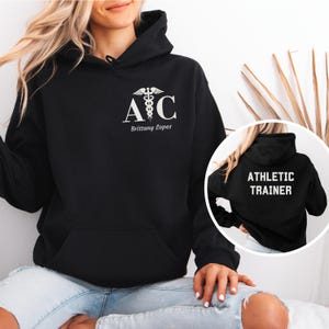 May include: Black hoodie with a white athletic trainer logo on the front and the text "Athletic Trainer" on the back. The logo features a caduceus, a symbol associated with medicine, and the letters "A" and "C". The name "Brittany Lopez" is printed below the logo.