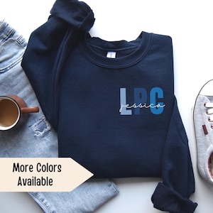 May include: A navy blue sweatshirt with the letters "LPC" in blue, white, and light blue shades. The name "Jessica" is written in white script below the letters. The sweatshirt is laid flat on a white surface with a pair of blue jeans and a gray shoe.