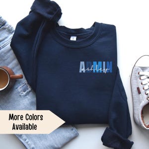 May include: A navy blue sweatshirt with the text "ADMIN ashley" in blue and white lettering. The sweatshirt is laid flat on a white surface with a pair of jeans and a pair of sneakers.