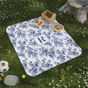 May include: A white and blue picnic blanket with a floral pattern and a navy blue monogram "R" in the center. The blanket is laid out on a grassy lawn.