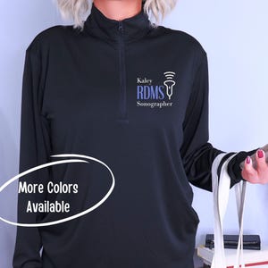 May include: Black mock neck shirt with a zipper and a blue logo that says "Kaley RDMS Sonographer" with a stylized ultrasound probe graphic. The text "More Colors Available" is written in a white circle.