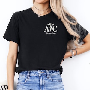 May include: A black short-sleeved t-shirt featuring a white graphic on the left chest. The design includes the letters A and C with a caduceus symbol between them and the name Brittany Lopez printed in white text underneath the letters.
