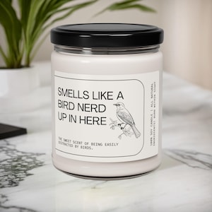 May include: A white soy candle with a black lid and a label that reads "Smells Like a Bird Nerd Up In Here." The label also includes a small illustration of a bird on a branch and the text "The sweet scent of being easily distracted by birds."