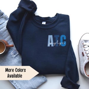 May include: A navy blue sweatshirt with the letters ATC in blue, white, and gray with the name Jaylyn written in white script underneath. The sweatshirt is laying on a white surface with a pair of blue jeans and a gray and white sneaker.