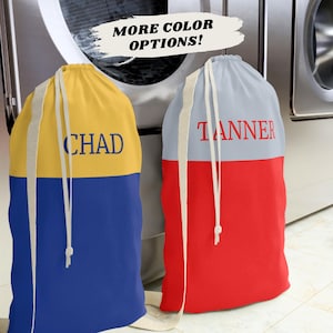 May include: Two personalized laundry bags. One is yellow and blue with "CHAD" in blue text. The other is red and gray with "TANNER" in red text. Both have white drawstring closures and straps. The text "MORE COLOR OPTIONS!" is above the bags.