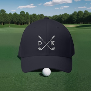 Peut inclure: Casquette de baseball bleu marine avec des clubs de golf blancs brodés qui se croisent. Les lettres "D" et "K" sont brodées sur la casquette. Une balle de golf blanche est devant la casquette sur une surface verte.
