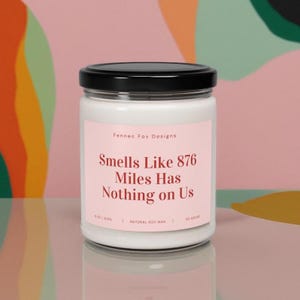 May include: A white candle with a black lid and a pink label that reads "Fennec Fox Designs Smells Like 876 Miles Has Nothing on Us 8 oz / 255g NATURAL SOY WAX 50 HOURS".
