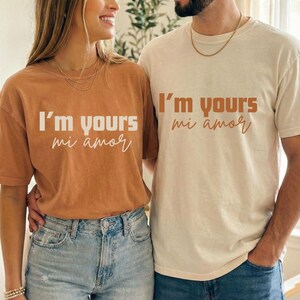 以下が含まれることがあります： 暖色系の茶色とクリーム色の2枚の半袖Tシャツには、対照的な色で「I'm yours mi amor」の文字がプリントされています。 シャツは青色のデニムジーンズと組み合わせています。 この画像は、カップルのペアの服を示唆しています。