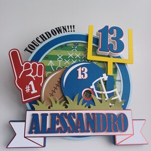 May include: A blue and white football themed cake topper with the name Alessandro and the number 13. The topper features a football helmet, a football, a goal post, and a hand with a number 1 on it. The words "Touchdown!!" are written on the top of the topper.