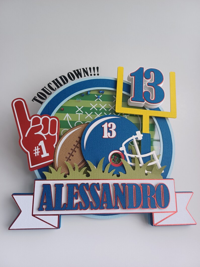 May include: A blue and white football themed cake topper with the name Alessandro and the number 13. The topper features a football helmet, a football, a goal post, and a hand with a number 1 on it. The words "Touchdown!!" are written on the top of the topper.