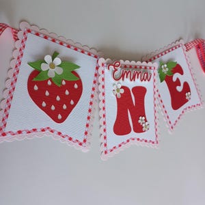 May include: A red and white banner with the name "Emma" and the number "One" in red letters. The banner is decorated with strawberries and white flowers.