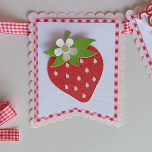 May include: A red and green paper strawberry with a white flower and green leaves. The strawberry is on a white background with a pink and red checkered border.