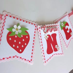 May include: A red and white gingham banner with the word "Emma" and the number "One" in red. The banner also features a strawberry with green leaves and a white flower.