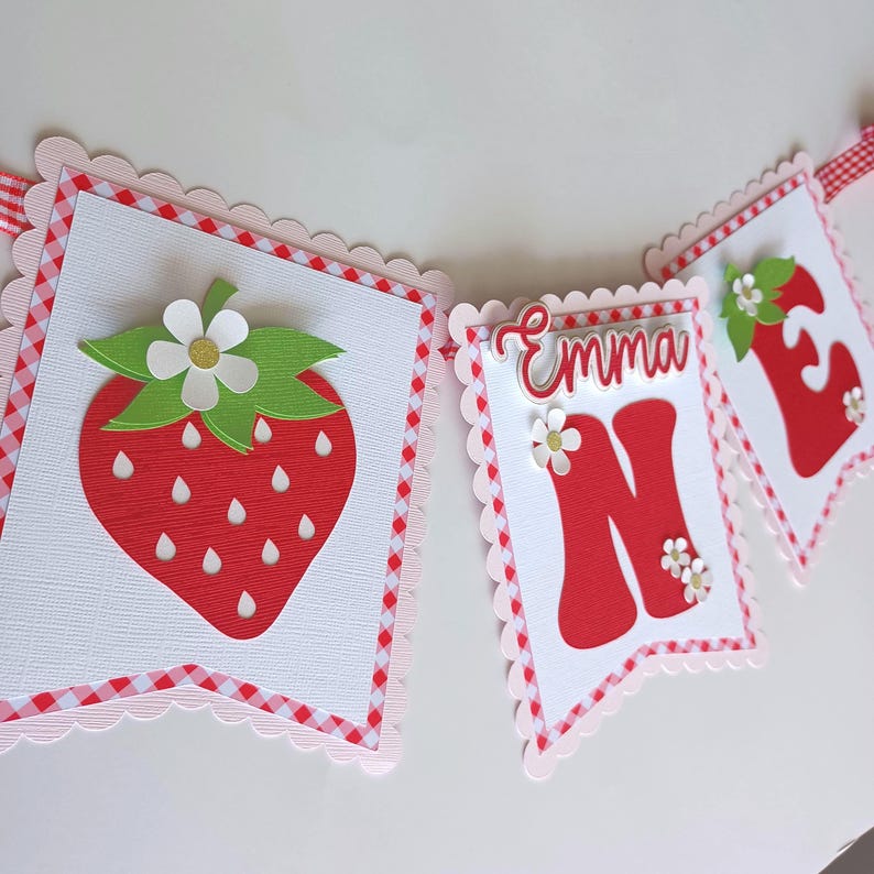 May include: A red and white gingham banner with the word "Emma" and the number "One" in red. The banner also features a strawberry with green leaves and a white flower.