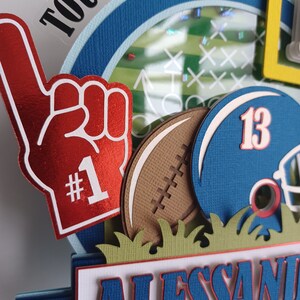 May include: A red and white foam finger with the number "#1" on it, a brown football, a blue football helmet with the number "13" on it, and a green grass border. The background is a blue and white circle with the word "TOUCHDOWN!!" in black letters.