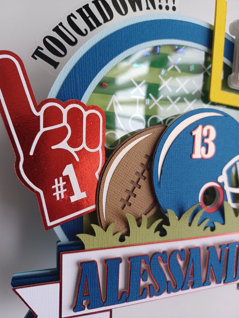 May include: A red and white foam finger with the number "#1" on it, a brown football, a blue football helmet with the number "13" on it, and a green grass border. The background is a blue and white circle with the word "TOUCHDOWN!!" in black letters.