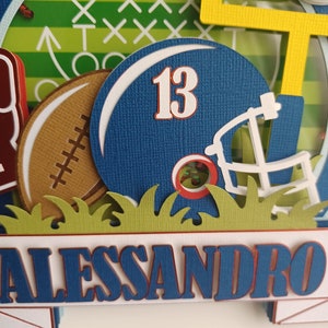 May include: A blue and white football helmet with the number 13 on it. The helmet is surrounded by green grass and a brown football. The name Alessandro is written in blue letters on a white banner.