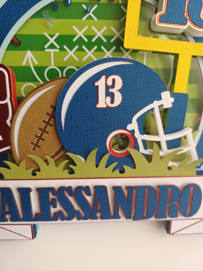 May include: A blue and white football helmet with the number 13 on it. The helmet is surrounded by green grass and a brown football. The name Alessandro is written in blue letters on a white banner.