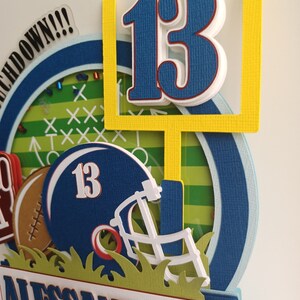 May include: A blue and yellow football themed card with the number 13, a football helmet, and the name Alessandro. The card has a green grass background and a white border.
