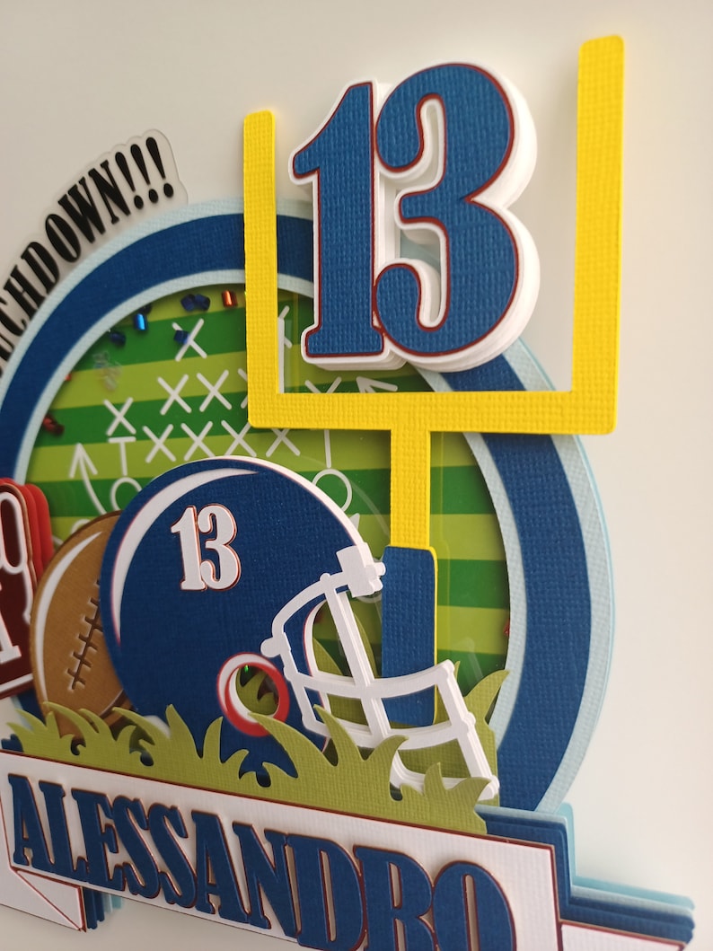 May include: A blue and yellow football themed card with the number 13, a football helmet, and the name Alessandro. The card has a green grass background and a white border.