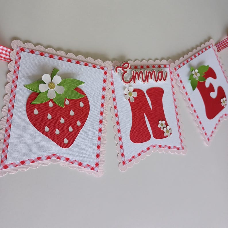 May include: A red and white banner with the name "Emma" and the number "One" in red letters. The banner is decorated with strawberries and flowers.