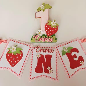 May include: A red and white banner with the name "Emma" and the number "one" decorated with strawberries and flowers. The banner is perfect for a first birthday party.