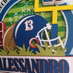 May include: A blue and red football helmet with the number 13 on it, a brown football, and a yellow goal post. The background is a green and white football field with the words "Touchdown!" in blue and white. The name "Alessandro" is written in blue and red on a white banner.