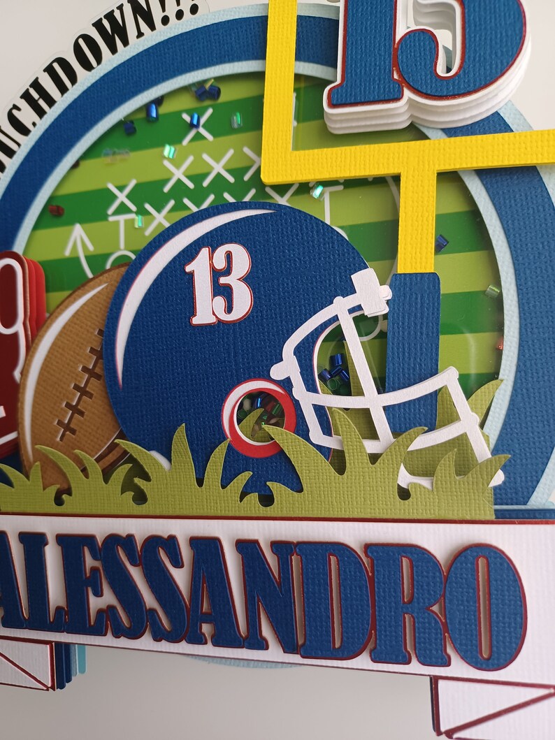 May include: A blue and red football helmet with the number 13 on it, a brown football, and a yellow goal post. The background is a green and white football field with the words "Touchdown!" in blue and white. The name "Alessandro" is written in blue and red on a white banner.