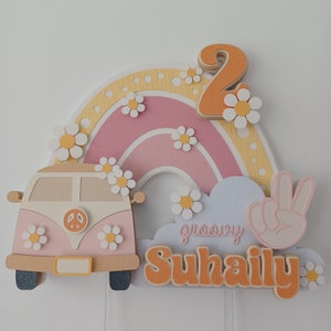 May include: A pink and yellow rainbow cake topper with a white camper van, a peace sign, and the text "groovy Suhaily" in orange. The number 2 is also included in orange.