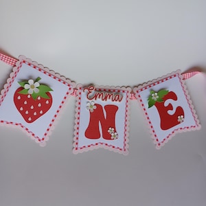 May include: A red and white banner with the name "Emma" and the number "one" in red letters. The banner is decorated with strawberries and flowers.