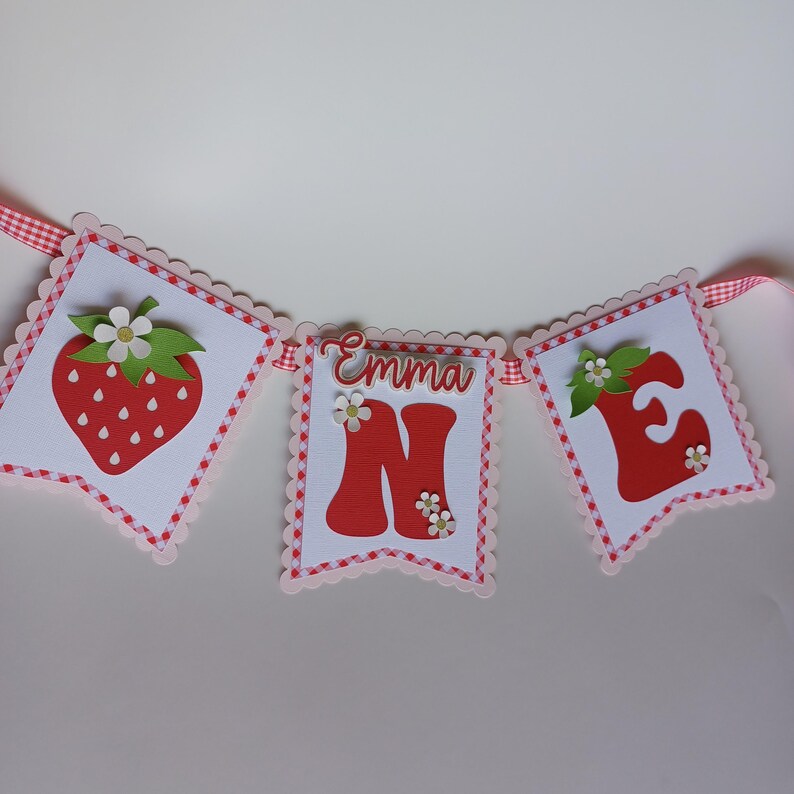 May include: A red and white banner with the name "Emma" and the number "one" in red letters. The banner is decorated with strawberries and flowers.