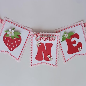 May include: A red and white banner with the word "Emma" and the number "one" in large red letters. The banner is decorated with strawberries and green leaves.