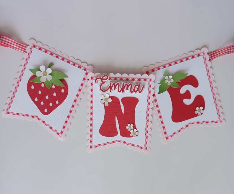 May include: A red and white banner with the word "Emma" and the number "one" in large red letters. The banner is decorated with strawberries and green leaves.