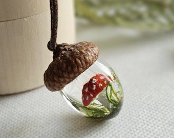 Op de afbeelding: Een glazen eikel hanger met een rode en witte paddenstoel erin. De eikel is gemaakt van hars en heeft een bruine dop. De hanger is aan een bruin koord.