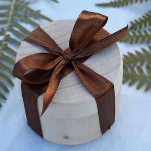May include: A round wooden box with a brown satin ribbon tied in a bow on top. The box is sitting on a white surface.