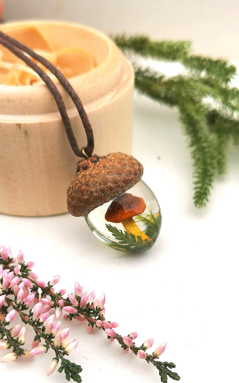 May include: A brown acorn pendant necklace with a clear resin dome containing a small orange mushroom and green foliage.