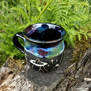 May include: A ceramic mug with a glossy, multi-colored glaze in shades of deep blue, purple, and teal at the rim, transitioning to a matte black base. The black section features etched white and gold celestial designs, including a planet and stars.