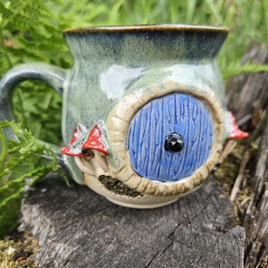 May include: A ceramic mug with a green glaze featuring a sculpted blue circular door with a black knob. The mug is decorated with small red and white mushrooms on the side and a textured stone-like frame around the door element.