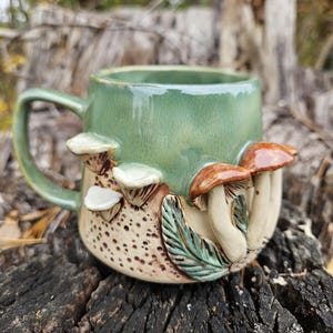 Puede incluir: Una taza de cerámica con una sección superior verde brillante y una base texturizada de color crema. El exterior está decorado con setas esculpidas en tres dimensiones en tonos marrones y blancos, junto con detalles de hojas verdes y puntos.