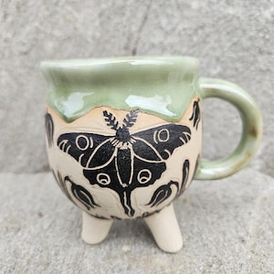 Puede incluir: Taza de cerámica hecha a mano con borde y asa verde claro. El cuerpo beige presenta un diseño de polilla negra y tres pequeños pies. La taza está diseñada para bebidas.