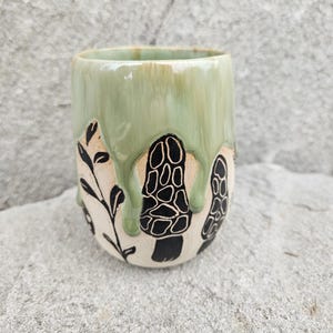 Può includere: Tazza in ceramica con una glassa verde chiaro che cola sui lati. La tazza presenta disegni di funghi e foglie neri. La tazza è cilindrica e sembra fatta a mano.