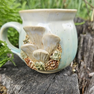 Könnte beinhalten: Ein Keramikbecher mit einer glänzenden, blassblauen und cremefarbenen Glasur. Die Außenseite zeigt eine dreidimensional geformte Ansammlung von Austernpilzen und texturierte moosartige Details in Braun-, Beige- und Grüntönen an der Seite.