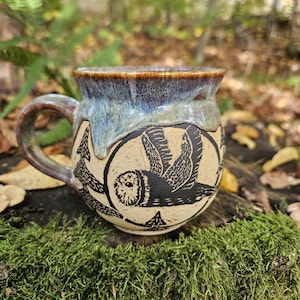 Peut inclure: Une tasse en céramique avec un corps arrondi et une anse incurvée. La partie supérieure présente un émail brillant bleu et marron, tandis que la partie inférieure est crème avec une illustration noire gravée d'un hibou en vol et de sapins.