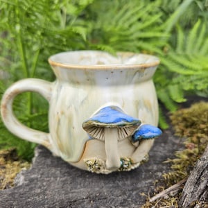 Op de afbeelding: Een keramische mok met een crèmekleurig glazuur, een druipend patroon en twee gebeeldhouwde, driedimensionale paddenstoelen aan de voorzijde. De paddenstoelen hebben felblauwe hoeden en lichte stelen, met kleine textuurdetails aan de onderkant.