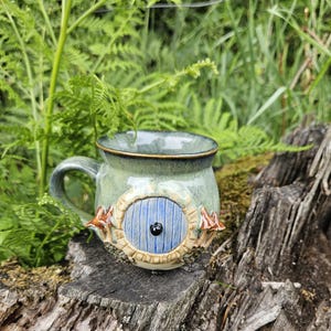 Pode incluir: Uma caneca de cerâmica com esmalte verde manchado, apresentando uma porta azul redonda esculpida com um puxador preto no centro. A porta é emoldurada por um arco bege texturizado, ladeada por pequenas figuras de cogumelos castanhos nos lados.